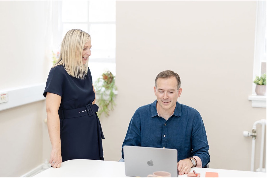 A woman stands smiling next to a man who is seated and working on a laptop at a white desk in a bright, modern office.
