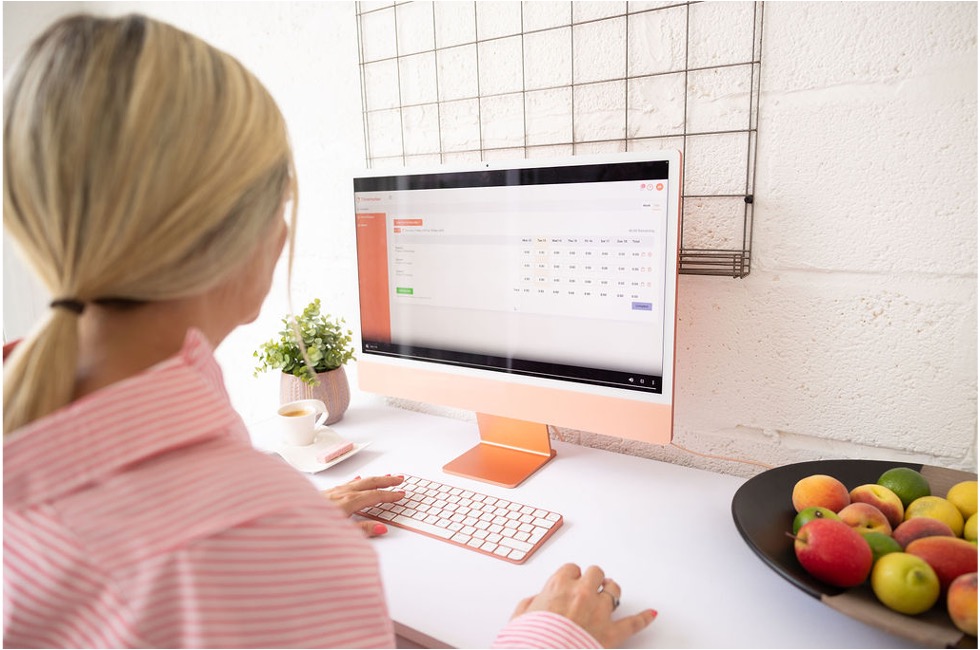 A woman with blonde hair in a pink striped shirt works on a desktop computer, with a bowl of fruit, a cup of coffee, and a potted plant nearby. The computer screen displays a Timesheet dashboard.