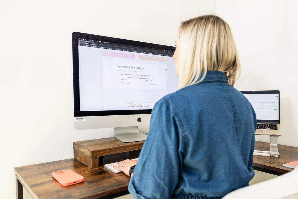 A person with blonde hair sits at a desk working on a computer using Timemarker, with a phone and a laptop