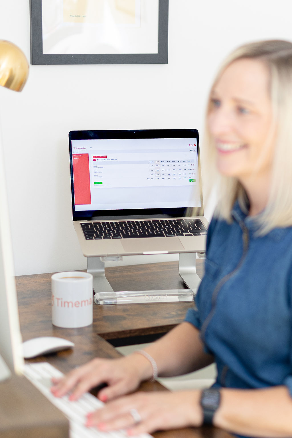 A person types on a desktop computer at a desk, with a Timemarker calendar open on the laptop in the background.