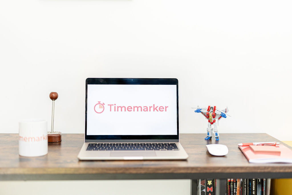 A laptop displaying the Timemarker logo sits on a wooden desk, surrounded by a mug, a retro stamp, a toy robot, a mouse, and some notebooks. The Timemarker logo stands out against the plain white wall in the background.
