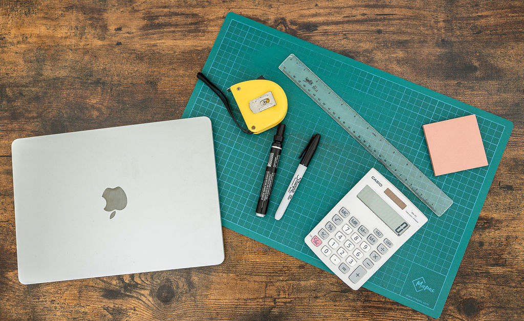 A closed silver MacBook, yellow tape measure, two markers, ruler, calculator, and pink sticky notes are neatly arranged with other stationary on a green cutting mat atop a wooden table.