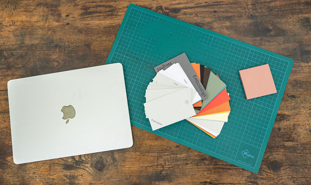 A silver MacBook, a green cutting mat, and a set of colourful paper swatches in the the brands pallet are arranged on a wooden table.