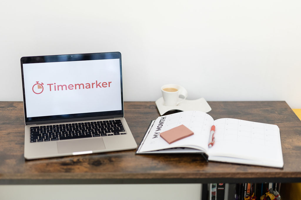 A laptop displaying Timemarker, an open monthly planner with a pen and sticky notes, and a cup of coffee on a wooden desk create an inspiring workspace for planning.