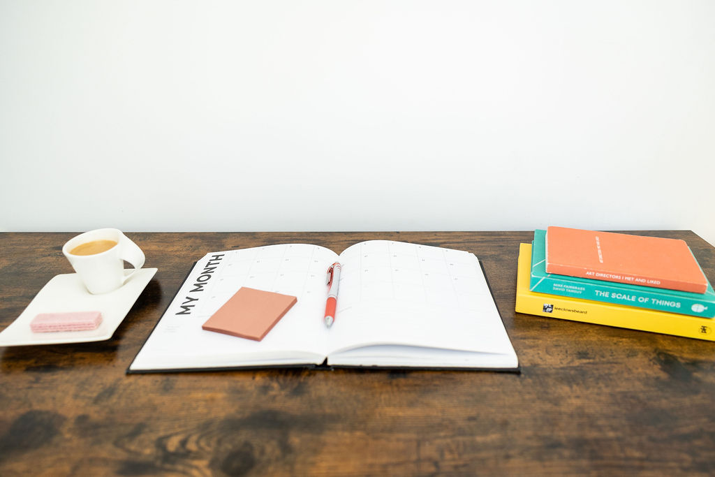A wooden desk with an open monthly planner for easy planning, a pen, a notepad, a cup of coffee with a biscuit, and a stack of colourful books.