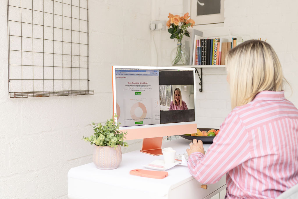A woman in pink sits at a desk, having a video call on her whilst using Timemarker on her computer. A plant, cup, and notebook are on the desk, with a shelf of books and flowers in the background.