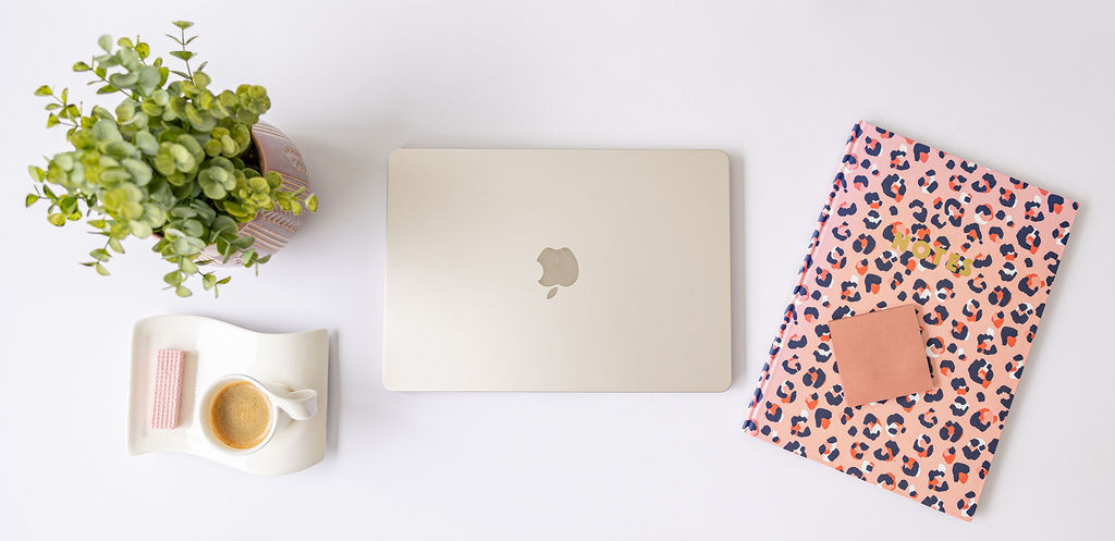 A silver MacBook, a leopard print notebook with a sticky note, a cup of espresso with wafers, and a potted green plant arranged on a white desk.