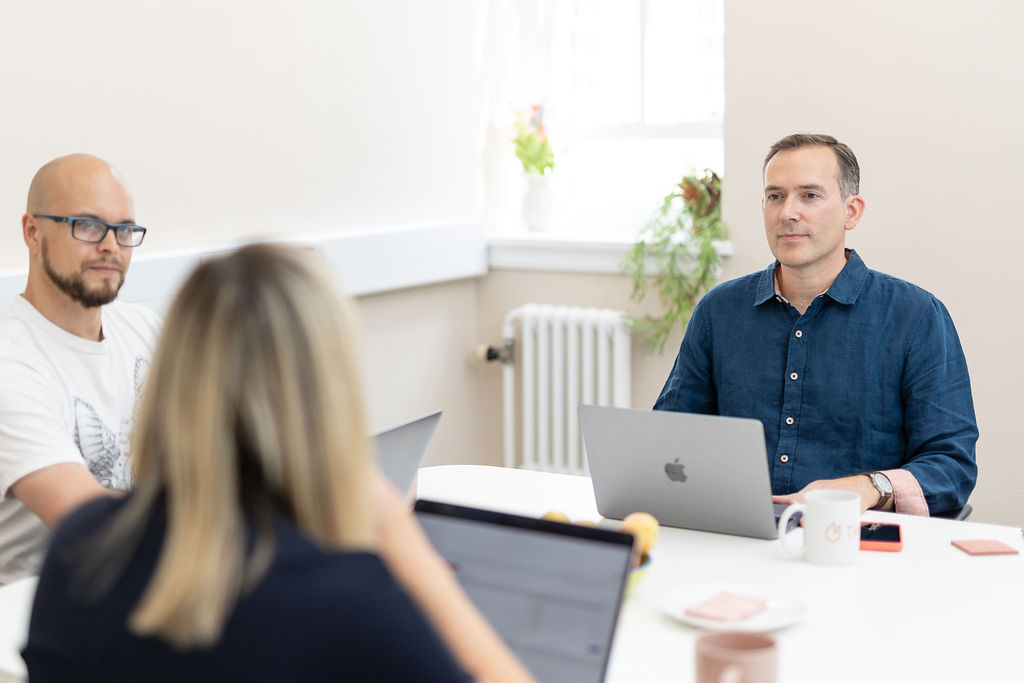 Three people sit at a table with laptops and coffee mugs, engaged in a meeting using Timemarker, in a bright office.