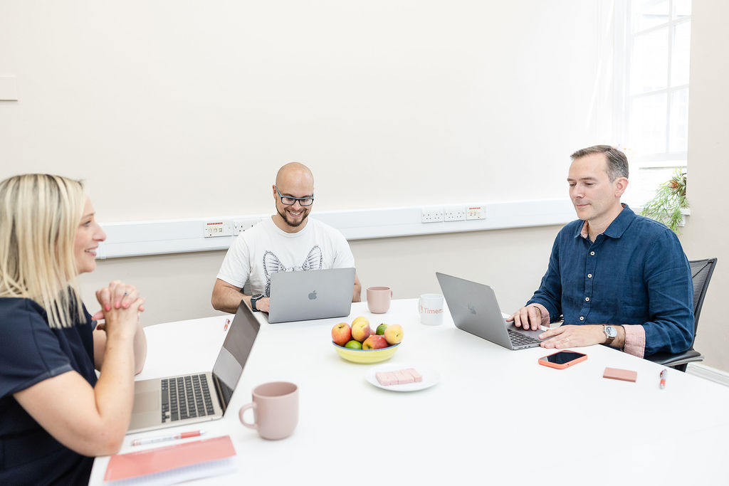 Three people sit around a white table with laptops, working. The table features fruit, mugs, notepads, and a Timemarker mug.. Natural light streams in through the window in the background.