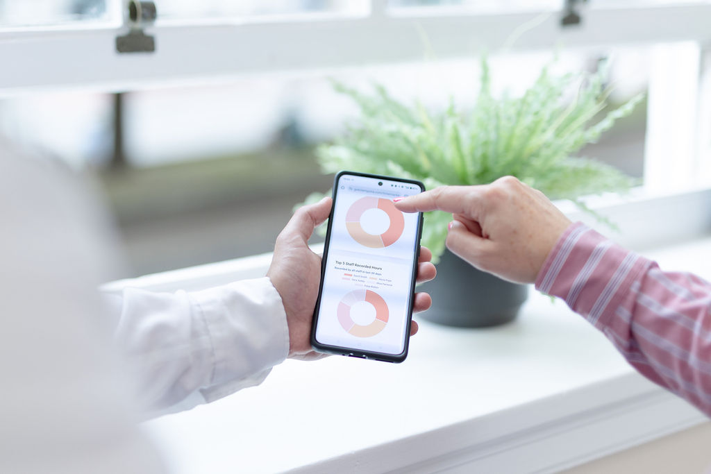 Two people view a smartphone displaying data visualisations with donut charts near a window. One person points at the top right of the screen, and there is a green potted plant in the background.
