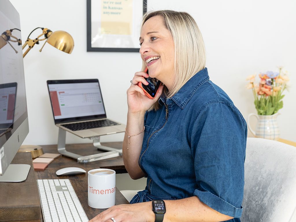A woman in a blue dress sits at a desk, smiling while talking on the phone. She works at a computer, with a, mug, and flowers nearby. A framed quote hangs on the wall behind her.