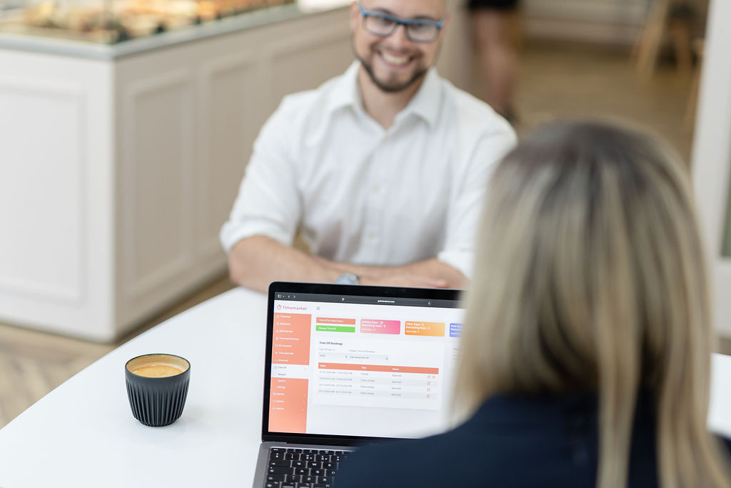 Two people sit at a table with a laptop and a coffee mug, engaged in a meeting using Timemarker, in a bright office.