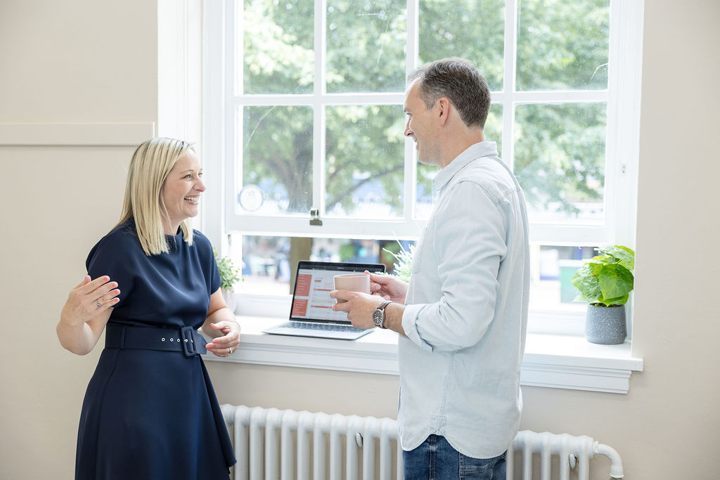 A woman stands smiling next to a man engaged into working on a laptop at a white windowsill in a bright, modern office.